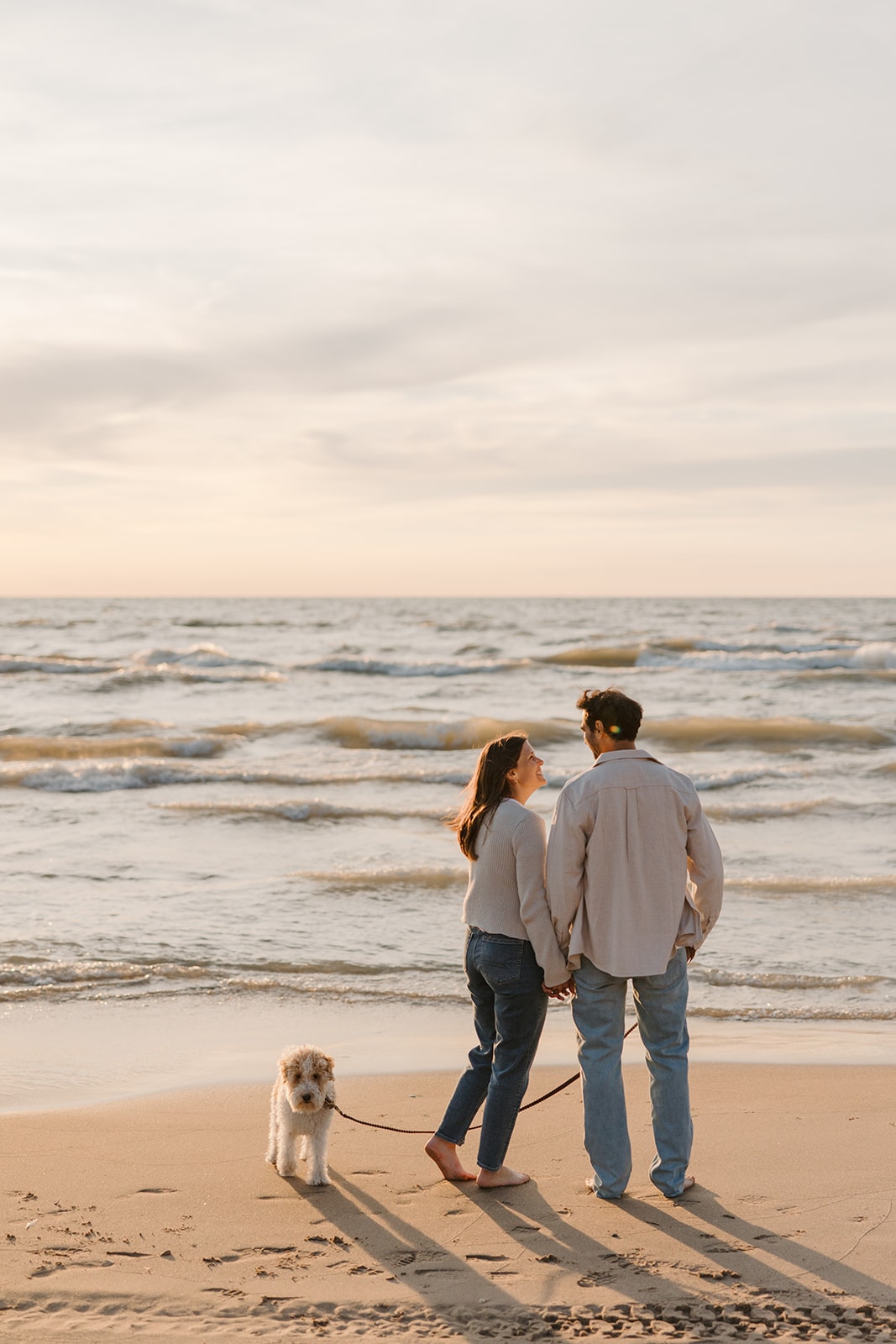Sarah and Stephane standing hand in hand on the beach beside their dog.